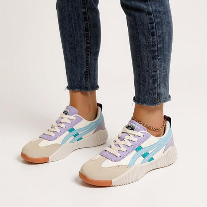 Damen tragen Onitsuka Tiger Sneaker aus Wildleder und Mesh in Beige, Weiß, Flieder und Hellblau mit gezackter Sohle, kombiniert mit dunkelblauen Jeans – modische und bequeme Freizeitschuhe